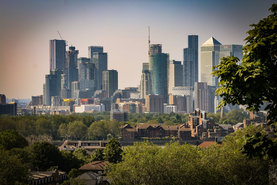 A panoramic view of a city skyline during daylight, featuring numerous modern high-rise office buildings and residential skyscrapers constructed with glass facades and steel frameworks. In the foregro