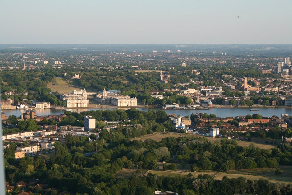 A wide-angle view of a large, historic white building with multiple wings and towers, situated in a spacious green park with neatly maintained lawns and pathways, in front of a modern city skyline com