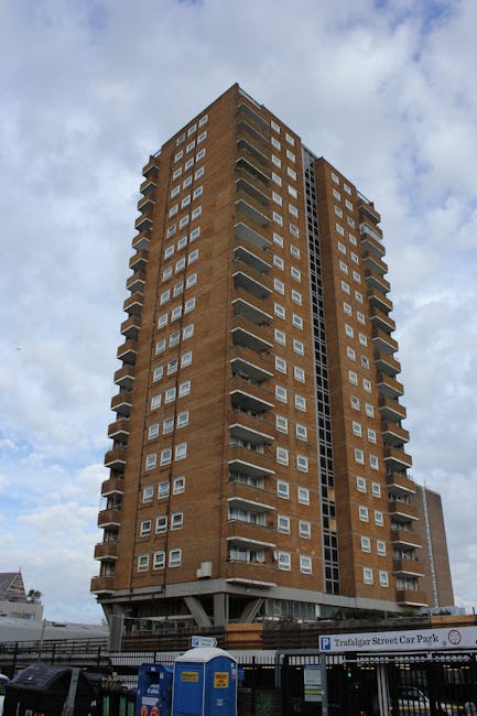 A tall, multi-storey residential building constructed with brown brickwork, featuring numerous white-framed windows and small balconies on each floor. The building is situated on a paved street, with